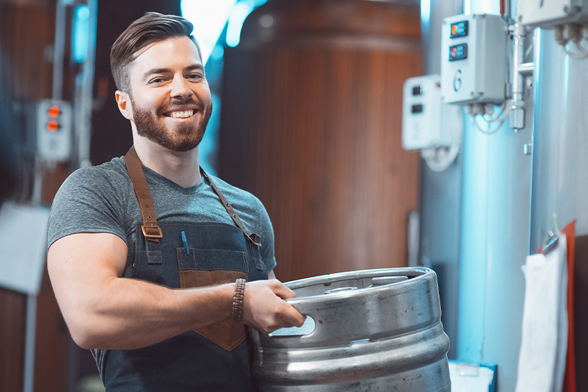 A young brewer in an apron holds a barrel with beer in the hands of a brewery. But how many beers in a keg?